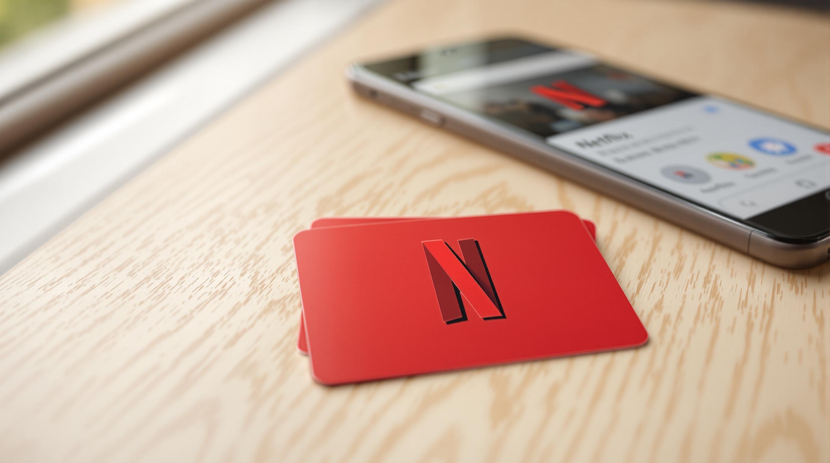 Red Netflix gift card on a wooden table, with a smartphone displaying the Netflix app in the background.