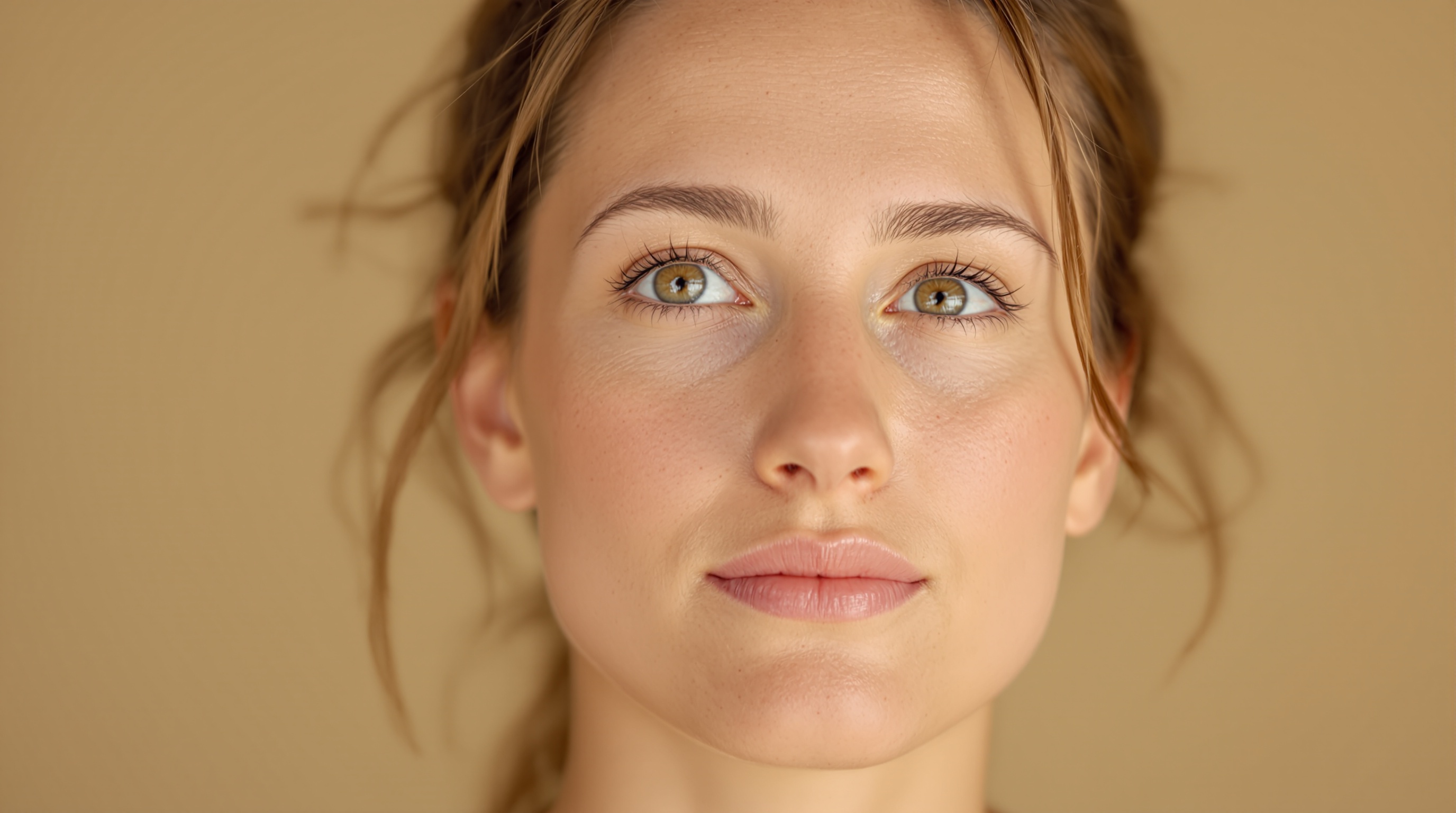 Portrait rapproché d'une femme au teint clair avec des yeux verts, des cheveux châtains attachés et un fond beige uni.