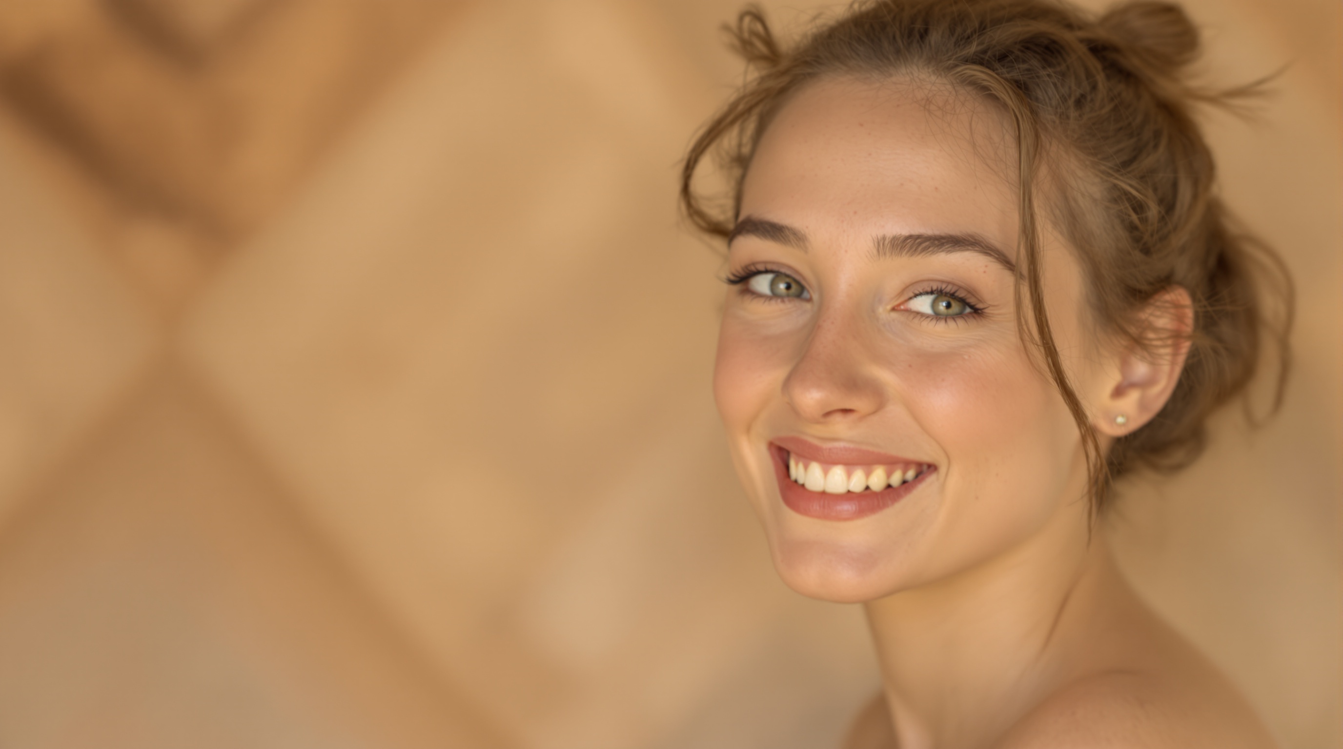 Femme souriante aux yeux clairs, cheveux attachés, devant un fond beige flou.