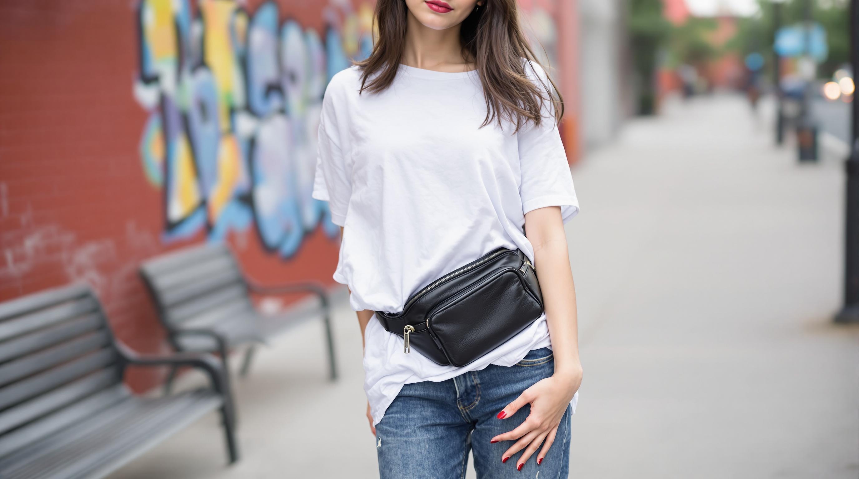 Femme portant un t-shirt blanc et un sac banane noir, debout dans une rue urbaine avec un mur de briques décoré de graffitis en arrière-plan.